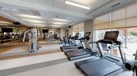 This fitness center is a spacious, single-floor facility designed to support a comprehensive workout experience. The space is thoughtfully laid out, with distinct zones for cardio and strength training that together create an organized, functional environment.

The cardio area features a row of treadmills and stationary bikes positioned along large windows fitted with roller shades, allowing ample natural light while offering adjustable privacy. The views to the outdoors contribute to an open, airy atmosphere throughout the space.

The strength training zone is equipped with multiple Cybex cable and weight machines, along with a dedicated ballet barre and full-length mirrored wall that visually expand the room and support a variety of exercise routines.

Underfoot, the flooring consists of durable, multi-toned vinyl tile in neutral grey, beige, and tan striped patterns, providing both visual interest and practical durability. Overhead, a suspended acoustic tile ceiling with integrated fluorescent lighting and ceiling fans ensures consistent illumination and comfortable air circulation throughout.

The space is finished with neutral grey walls, wood-toned accents, and clean lines, creating a modern yet welcoming fitness environment suited for a wide range of users.
