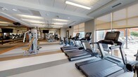 This fitness center is a spacious, single-floor facility designed to support a comprehensive workout experience. The space is thoughtfully laid out, with distinct zones for cardio and strength training that together create an organized, functional environment.

The cardio area features a row of treadmills and stationary bikes positioned along large windows fitted with roller shades, allowing ample natural light while offering adjustable privacy. The views to the outdoors contribute to an open, airy atmosphere throughout the space.

The strength training zone is equipped with multiple Cybex cable and weight machines, along with a dedicated ballet barre and full-length mirrored wall that visually expand the room and support a variety of exercise routines.

Underfoot, the flooring consists of durable, multi-toned vinyl tile in neutral grey, beige, and tan striped patterns, providing both visual interest and practical durability. Overhead, a suspended acoustic tile ceiling with integrated fluorescent lighting and ceiling fans ensures consistent illumination and comfortable air circulation throughout.

The space is finished with neutral grey walls, wood-toned accents, and clean lines, creating a modern yet welcoming fitness environment suited for a wide range of users.