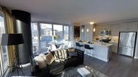 This contemporary single-floor residence showcases a thoughtfully designed open-concept layout that seamlessly integrates the living, dining, and kitchen areas into one cohesive, light-filled space. Floor-to-ceiling windows dominate the living area, flooding the interior with natural light and framing expansive views of the surrounding urban skyline. The living room centers around a bold leather sofa accented with decorative throw pillows and a textured area rug, creating a sophisticated yet comfortable atmosphere. Adjacent to the living space, a compact dining area with a round table and modern chairs sits directly beside the windows, making it an ideal spot to enjoy city views. The kitchen is sleek and modern, featuring light cabinetry, stainless steel appliances, and a marble-topped peninsula with bar seating — perfect for casual dining or entertaining. Pendant lighting adds warmth and visual interest above both the kitchen peninsula and dining area. Dark hardwood floors run throughout, providing a rich contrast to the bright, neutral walls and cabinetry. Track lighting on the ceiling further enhances the contemporary aesthetic. Overall, the space exudes a refined urban ambiance, blending modern finishes with an inviting, functional design.