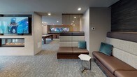 This contemporary club room is thoughtfully designed to accommodate a wide range of social activities within a single, well-appointed space. The layout flows seamlessly between distinct zones, each serving a unique function while maintaining a cohesive modern aesthetic throughout.

The kitchen area is a standout feature, anchored by a large granite-topped island with an integrated cooktop and overhead range hood. Stainless steel appliances, a textured stone tile backsplash, and warm hardwood flooring give the kitchen a polished yet inviting character. Bar-height seating along the island and adjacent windows allow natural light to fill the space.

Adjacent to the kitchen, a comfortable lounge area features a generous sectional sofa in teal and charcoal, centered around a reclaimed wood coffee table beneath a distinctive geometric pendant light. Full-height glass doors open directly to an outdoor terrace, blending interior and exterior entertaining spaces.

The games area is well-equipped with a full-size billiards table, a shuffleboard table, and a linear fireplace set against a textured stone accent wall. Cove lighting along the ceiling adds a warm, ambient glow. A secondary lounge zone offers high-back seating, mounted flat-screen televisions, and a translucent room divider, providing a more intimate setting within the larger space.