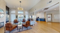 This single-floor community clubhouse, known as The Villas, is thoughtfully designed to provide residents with an inviting and well-appointed common area. The main lounge is the heart of the space, boasting soaring vaulted ceilings, light wood-look flooring, and an abundance of natural light streaming through large windows dressed with sheer white curtains. A striking modern chandelier anchors the seating area, which features rich cognac leather armchairs, a plush sofa, and patterned area rugs arranged around a contemporary fireplace set against a floor-to-ceiling stone surround. Bold abstract artwork adorns the accent walls, adding a vibrant artistic touch throughout.

A dedicated refreshment bar includes navy blue cabinetry, a beverage refrigerator, a microwave, a sink, and a coffee station, all set against a glossy white subway tile backsplash. Adjacent to the lounge is a communal dining area with a natural wood farmhouse-style table positioned near oversized windows with outdoor views.

A separate lounge room offers additional seating with bold orange accent chairs, a large-screen television, and vibrant wall art. The fitness center is well-equipped with treadmills, stationary bikes, and strength training equipment, and features large windows overlooking the outdoor grounds with views of palm trees.