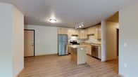 This single-floor space offers a clean, functional layout centered around an open-concept kitchen and living area. The kitchen is well-appointed with light maple cabinetry extending to the ceiling, providing generous storage throughout. Stainless steel appliances — including a full-size refrigerator, electric range, and dishwasher — lend a modern, cohesive look to the space. A central kitchen island adds additional prep space and serves as a natural dividing point between the kitchen and the open living area beyond. Granite-style countertops complement the warm wood tones of the cabinetry, while a stainless steel sink is positioned beneath a dedicated under-cabinet workspace along the back wall. Track lighting above the kitchen provides focused illumination, supplemented by a flush-mount ceiling fixture that brightens the broader living space. Light hardwood-style flooring runs seamlessly throughout the entire unit, enhancing the sense of openness and continuity. Neutral-toned walls keep the interior feeling bright and versatile. The entryway, visible from the kitchen, features a solid interior door, offering a welcoming and practical transition into the living space. Overall, the unit presents a tidy, move-in-ready environment with a warm and approachable aesthetic.