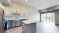 This single-floor residence offers a thoughtfully designed open-concept layout that seamlessly connects the kitchen and living areas. The kitchen is a standout feature, showcasing two-tone cabinetry with crisp white upper cabinets and grey lower cabinets, light quartz countertops, and a full suite of stainless steel appliances including a refrigerator, range, microwave, and dishwasher. A central island provides additional prep space and serves as a natural gathering point. Vaulted ceilings in the kitchen and living area lend an airy, expansive feel to the space, while light-toned luxury vinyl plank flooring runs throughout the main living areas. A sliding glass door opens to a private balcony, allowing natural light to flood the interior. The bedrooms are carpeted for comfort and feature generous proportions with windows that bring in outdoor views. Each bedroom is complemented by walk-in closet access. The bathroom is clean and functional, featuring a full tub-shower combination with chrome fixtures and wood-look flooring. A dedicated in-unit laundry room with a full-size washer and dryer adds everyday convenience. Neutral tones and recessed lighting throughout create a bright, contemporary atmosphere.