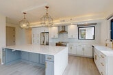 This single-floor space showcases a cohesive modern design defined by a crisp white-and-gold palette, light-toned large-format tile flooring, and an abundance of natural light. The heart of the space is a generously proportioned open-plan kitchen and living area. The kitchen is outfitted with floor-to-ceiling white shaker cabinetry adorned with brushed gold hardware, a substantial central island featuring a contrasting slate-blue base topped with a thick white quartz surface, and a matching quartz backsplash surrounding the professional-grade range beneath a stainless steel hood. A farmhouse sink with a gold faucet sits beneath black-framed windows that frame lush outdoor greenery. Geometric gold pendant lights hang overhead, adding a sculptural element to the space. Stainless steel appliances, including a refrigerator, dishwasher, and built-in microwave, complete the culinary suite. A dedicated laundry closet houses a stacked washer and dryer. The living area is expansive and bright, bordered by floor-to-ceiling black-framed sliding glass doors and windows that open to a balcony with verdant views. The bathroom features floor-to-ceiling marble-look tile in the walk-in glass-enclosed shower, paired with a blue-grey vanity, round mirror, and gold fixtures for a polished, spa-like finish.