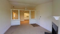 This single-floor space offers an open and airy layout defined by soaring vaulted ceilings that lend a sense of spaciousness and grandeur. The living area is anchored by a ceiling fan and flows effortlessly into the kitchen, separated by a pair of decorative white columns and a half-wall with built-in shelving — a charming architectural detail that adds both character and practicality. The built-in shelves provide convenient display and storage space while maintaining visual openness between rooms.

The kitchen is bright and well-appointed, featuring white cabinetry, under-cabinet lighting, and white appliances. A window above the sink allows natural light to filter in, keeping the space feeling fresh and inviting. The transition between rooms is marked by a shift from soft, neutral carpeting in the living area to hardwood-style flooring near the entryway and kitchen, adding warmth and texture throughout.

The entryway is positioned conveniently beside the living space, framed by a clean white door. The overall color palette — soft whites, warm neutrals, and light wood tones — creates a cohesive and welcoming ambiance throughout the home, making it a bright, functional, and comfortable space.