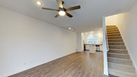 This 2-floor space offers a clean, modern layout with an open-concept main level that seamlessly connects the living area and kitchen. The living area is finished with warm hardwood-style plank flooring that extends throughout, complemented by crisp white walls that create a bright, airy atmosphere. A ceiling fan with an integrated light fixture anchors the living space, while recessed track lighting overhead provides additional illumination. The open kitchen is well-appointed with white cabinetry, a center island, stainless steel appliances including a dishwasher and refrigerator, and a window that allows natural light to filter in, offering views of the surrounding greenery. The kitchen's layout maximizes functionality while maintaining visual flow with the living area. A partial white column and staircase detail suggest a thoughtfully designed transition to the second floor. The overall aesthetic is contemporary and neutral, making it a versatile and move-in-ready space suited to a variety of lifestyles.