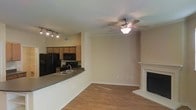 This two-story space features an open-concept main living area that seamlessly connects the kitchen and living room. The living room is anchored by a white-mantled fireplace and is equipped with a ceiling fan with integrated lighting, complemented by crown molding that adds a refined architectural touch. Hardwood-style flooring runs throughout, providing a warm and cohesive aesthetic.

The kitchen is positioned adjacent to the living area and is defined by a raised breakfast bar with a dark countertop, offering both a functional workspace and a casual dining option. Cabinetry in a warm wood tone provides ample storage, while black appliances, including a refrigerator and microwave, add a modern contrast. A double sink is conveniently located along the bar counter. Track lighting above the kitchen ensures the workspace is well-lit.

Neutral-toned walls throughout the main level create a clean, versatile backdrop suitable for a variety of interior styles. The open layout promotes a sense of spaciousness, while the overall design balances practicality with comfortable, everyday living across both floors.