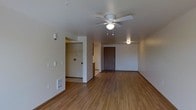 This single-floor space offers a clean, functional layout designed for comfortable everyday living. The main living area is anchored by warm hardwood-style flooring that extends throughout, creating a cohesive and inviting atmosphere. A ceiling fan with an integrated light fixture is centrally mounted, providing both air circulation and ambient lighting to the space.

The living area flows naturally toward the front entry, which features a solid dark-toned door that adds a grounding contrast to the neutral, light-colored walls. A floor vent ensures climate control is well-integrated into the design. Adjacent to the main living space, an open passage leads to what appears to be a utility or laundry alcove, partially visible and equipped with hookups, as well as access to additional areas of the unit.

The walls throughout are finished in a soft, neutral tone that reflects light well and complements the warm flooring beneath. The overall ambiance is bright, understated, and move-in ready, with a straightforward layout that makes the most of the available space. The open sightlines between areas enhance the sense of spaciousness, making this a practical and welcoming single-level home.