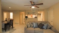 This single-floor space offers a warm and inviting atmosphere highlighted by neutral beige walls and light-toned carpeting throughout. The living area is the focal point of the home, centered around a generously sized sofa adorned with decorative pillows in complementary patterns and colors. A ceiling fan with a built-in light fixture is mounted overhead, providing both airflow and ambient lighting to the room. A decorative metal wall accent adds a subtle artistic touch above the pass-through opening that connects the living area to the kitchen.

The kitchen, visible through the pass-through counter, features warm wood-toned cabinetry and a white refrigerator, contributing to the home's cohesive color palette. The open sightlines between the kitchen and living area create a connected, sociable feel. A hallway visible from the living room leads to additional areas of the home, suggesting a functional and well-organized layout. Wall vents and floor registers indicate a central HVAC system throughout. The overall ambiance is comfortable and lived-in, with a traditional residential feel suitable for relaxed everyday living.