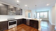This single-floor, 2-bedroom, 2-bathroom residence offers a thoughtfully designed layout that balances style and practicality. The open-concept kitchen serves as the heart of the home, showcasing dark espresso cabinetry, white quartz countertops, stainless steel appliances — including a range, microwave, and dishwasher — and a peninsula with a built-in sink and matte black faucet. Sleek pendant lighting accents the space, while rich hardwood flooring extends throughout the main living areas, adding warmth and continuity.

The living area is bathed in natural light from large windows and a glass door that opens to a private balcony, offering outdoor views. Louvered interior doors provide a clean, consistent aesthetic throughout the hallways, complementing the neutral wall tones.

Both bedrooms are carpeted and feature windows that bring in ample natural light. The primary bedroom includes a dedicated walk-in closet with built-in shelving and rod systems. The en-suite bathroom is equipped with a glass-enclosed walk-in shower featuring white tile surround and matte black hardware, with direct access to the walk-in closet. A second full bathroom and an additional carpeted closet serve the second bedroom. The unit also includes an in-unit laundry area accessible via louvered doors.
