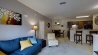 This 2-bedroom, 2-bathroom residence is thoughtfully designed on a single level, offering an efficient and comfortable open-concept floor plan. The living area features plush carpeted flooring and neutral gray walls that create a clean, modern backdrop, with ample space for seating and relaxation. The layout flows seamlessly into a dedicated dining area, illuminated by a stylish pendant light fixture that adds a warm, inviting focal point to the space. Adjacent to the dining area, the kitchen is appointed with rich dark cabinetry, a tile backsplash, and a breakfast bar with counter-height seating — ideal for casual dining or entertaining. The kitchen's hard-surface flooring provides a practical contrast to the carpeted living spaces. Ceiling-mounted vents ensure consistent climate control throughout. The open sightlines between the living, dining, and kitchen areas enhance the sense of spaciousness and connectivity. With two full bathrooms and two bedrooms, this residence offers both comfort and functionality, making it well-suited for a variety of living arrangements. The cohesive neutral color palette and contemporary finishes throughout give the space a polished, move-in-ready feel.