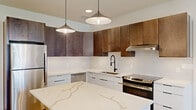 This contemporary single-floor unit offers a thoughtfully designed layout across five rooms, blending modern aesthetics with practical functionality. The kitchen serves as the centerpiece of the space, showcasing a striking waterfall-edge quartz island with bold veining, complemented by two-tone cabinetry — rich walnut uppers paired with crisp white lower cabinets. A white subway tile backsplash, matte black fixtures, and stainless steel appliances, including a refrigerator, dishwasher, and range, complete the polished look. Stylish pendant lights hang above the island, adding warmth and visual interest. Light-toned hardwood flooring flows seamlessly throughout the entire unit, unifying each space with a clean, cohesive feel. The bedroom area benefits from generous natural light streaming through large windows fitted with blinds, offering views of the surrounding cityscape and greenery. A dedicated laundry area houses a full-size washer and dryer, providing added convenience. The bathroom features a white vanity with quartz countertop, a framed mirror, matte black hardware, and accessibility grab bars. Additional rooms offer versatile, open spaces with neutral white walls and recessed lighting, ready to be personalized to suit a variety of uses.