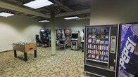 This single-floor arcade and game room offers a fun and engaging environment designed for entertainment. The space features an open layout with a patterned carpet floor and an exposed industrial-style ceiling supported by structural columns, giving it a casual, no-frills atmosphere well-suited to its purpose.

The room is equipped with a variety of arcade and gaming machines, including classic upright arcade cabinets, a modern racing or motion simulator chair setup, and at least one pinball machine, catering to a wide range of gaming preferences. The machines are arranged throughout the space, allowing for comfortable access and movement between stations.

Overhead, practical fluorescent and track lighting fixtures provide ample illumination across the gaming area, ensuring good visibility for players. A fully stocked snack vending machine is conveniently positioned near the entrance, offering a selection of packaged snacks for guests.

The overall ambiance is casual and recreational, reminiscent of a classic arcade hall. The space appears well-suited as a shared amenity or entertainment venue, providing guests or residents with a dedicated area for gaming and leisure.