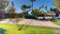Located at 725 S Bixel St in downtown Los Angeles, this well-appointed outdoor recreational space offers a variety of amenities set within a lush, tropical landscape. The grounds feature multiple hard-surface tennis courts with regulation nets, enclosed by green windscreen fencing and framed by soaring palm trees that provide natural shade throughout the day. Adjacent to the tennis courts, a sand volleyball court offers an additional recreational option, complete with a net and clearly defined boundaries bordered by manicured lawn. A rubberized walking path winds through the property, connecting the various activity areas while passing beneath mature trees and past richly planted garden beds filled with tropical foliage, succulents, and ornamental grasses. Classic lamppost fixtures line the pathway, adding a charming and functional lighting element to the grounds. Open lawn areas provide generous green space throughout, offering a relaxed, park-like setting. The entire space enjoys abundant natural sunlight under Southern California's clear blue skies, while striking views of the downtown Los Angeles skyline, including prominent glass high-rises, create a dramatic urban backdrop. The combination of active sport facilities, verdant landscaping, and city views makes this outdoor space a distinctive recreational destination in the heart of Los Angeles.