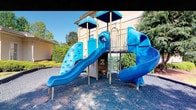 This outdoor playground is a well-equipped recreational space designed to engage and inspire active play. At its centerpiece stands a multi-level play structure finished in bold blue and neutral tan tones, offering a variety of features that cater to children of different ages and abilities. The structure includes two covered platform towers that provide shade and a sense of adventure, connected by walkways and open passages that encourage exploration. Two blue slides of varying configurations — including a straight run and a curved descent — offer exciting ways to navigate from the upper levels to the ground. A textured climbing wall panel with circular handholds provides a physical challenge, promoting strength and coordination. The ground beneath the structure is covered in rubber mulch, offering a safe, impact-absorbing surface. The play area is bordered by mature hedges and trees, creating a naturally enclosed and sheltered environment. A building with a light-colored exterior is visible adjacent to the space, suggesting the playground is part of a larger facility. Under open skies, the space is well-lit by natural sunlight, making it an inviting and energetic outdoor destination.