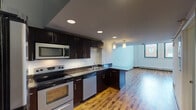 This contemporary two-floor residence showcases a thoughtfully designed open-concept layout that seamlessly connects the kitchen and living areas. The kitchen is a standout feature, outfitted with rich dark-espresso cabinetry, granite countertops, and a full suite of stainless steel appliances including an electric range, built-in microwave, dishwasher, and refrigerator. Recessed lighting and pendant fixtures illuminate the space, while warm hardwood flooring flows throughout the main living areas, adding a sense of richness and continuity.

The living area benefits from soaring double-height ceilings and expansive windows that invite abundant natural light and provide open views of the surrounding neighborhood. An interior mezzanine-style upper level overlooks the main floor, enhancing the sense of vertical space and architectural character. The upper level features carpet flooring and a dedicated closet area with a clothing rail.

The bathrooms are clean and functional, featuring wood-toned vanities with dark cabinetry, tub-shower combinations, and built-in shelving for added storage. A second half-bath with hardwood flooring provides added convenience. Throughout the space, the consistent use of wood tones, neutral walls, and modern fixtures creates a cohesive and inviting atmosphere.