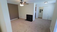 This single-story space features an open living area finished with wall-to-wall carpeting in a warm neutral tone, creating a comfortable and understated atmosphere. A decorative fireplace with a black arched screen serves as a focal point along the main wall, adding a classic touch to the room. A ceiling fan with an integrated light fixture is centrally mounted overhead, providing both air circulation and ambient lighting. The walls are painted in light beige with one accent wall in a deeper brown tone, offering subtle visual contrast. Adjacent to the living area, a passageway leads to what appears to be a kitchen, where dark cabinetry is partially visible, suggesting a functional and defined cooking space. A utility or electrical panel is visible through a nearby doorway, indicating practical infrastructure built into the layout. The open sightlines between rooms contribute to an easy flow throughout the space. Overall, the interior presents a clean, move-in-ready environment with a straightforward, functional design suited for comfortable everyday living.