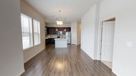 This single-floor unit showcases an open-concept layout that seamlessly connects the living area to the kitchen, creating a spacious and functional flow. The kitchen is outfitted with rich, dark espresso cabinetry contrasted by a white center island that doubles as a prep and gathering space. Stainless steel appliances, including a refrigerator, range, and microwave, complement the modern aesthetic, while a mosaic tile backsplash adds a decorative touch. Pendant lighting hangs above the island, providing focused illumination and a stylish focal point. The living area extends generously from the kitchen, offering ample room for furnishings and entertaining. Wide-plank, hardwood-style flooring runs throughout the main living space, adding warmth and continuity. Natural light enters through multiple windows fitted with horizontal blinds, brightening the neutral, light-toned walls. A doorway off the main space leads to additional rooms, suggesting a practical and well-organized floor plan. The overall ambiance is clean, contemporary, and move-in ready, with finishes that balance style and durability.