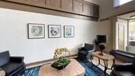This single-floor lobby presents a refined and inviting entrance experience defined by its dramatic double-height ceilings and a generous wall of clerestory windows above the main entry doors, flooding the space with natural light. Large-format neutral tile flooring extends throughout, lending a clean and polished aesthetic to the open floor plan.

The seating area is thoughtfully appointed with leather armchairs and a sofa arranged around a stone-topped accent table, anchored by a vibrant blue-toned area rug that adds a pop of color. An elegant urn-style lamp serves as a striking centerpiece. The walls are adorned with a series of framed nautical-inspired artworks, complemented by textured white walls and warm wood-paneled detailing along the upper mezzanine-level fascia.

A decorative console table featuring a bold ornate mirror flanked by sculptural candlestick holders adds a formal touch near an interior doorway that leads to a mailroom area. Tall potted greenery near the elevator bank introduces a natural element, enhancing the welcoming atmosphere. The main entry vestibule includes a kiosk station and bench seating, offering practical functionality alongside the lobby's polished design. Overall, the space balances professional sophistication with approachable warmth.