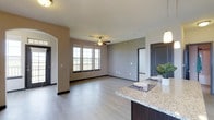 This single-floor space offers a thoughtfully designed open-concept layout that seamlessly connects the kitchen and living areas. The kitchen features a granite countertop island that serves as both a functional workspace and a casual dining area, complemented by dark cabinetry and stylish pendant lighting overhead. The expansive living area is finished with durable hardwood-style flooring and painted in warm neutral tones, creating a welcoming and versatile environment. A ceiling fan centered in the living space adds both comfort and a classic aesthetic touch. Natural light pours in through large double-hung windows framed in dark trim, offering views to the outdoors and enhancing the airy feel of the room. The layout includes clearly defined entryways and doorways with dark-painted doors, providing a modern contrast against the lighter wall tones. Additional rooms are accessible from the main living area, suggesting a practical and well-organized floor plan. Overall, the space combines contemporary finishes with functional design, making it an inviting and comfortable environment suited for everyday living.
