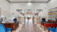 Located at 3501 E Gore Blvd in Lawton, Oklahoma, this single-floor community clubhouse offers a thoughtfully designed layout across a generous footprint. The reception and office area makes an immediate impression with its soaring tray ceiling, vibrant painted ceiling mural, and a striking yellow pendant light as a centerpiece. Built-in shelving with warm wood cabinetry lines the walls, while large floor-to-ceiling windows and glass entry doors flood the space with natural light and offer views of the surrounding outdoors. Light tile flooring and rich wood-toned desks with blue upholstered chairs complete the professional yet welcoming atmosphere. The adjoining living room is anchored by a full-height natural stone fireplace with a mounted television above, a coffered tray ceiling, and direct outdoor access through French doors that frame lush green exterior views. Built-in bookshelves add both function and character. The open kitchen and living area features rich wood cabinetry, stainless steel appliances including a double wall oven and French door refrigerator, granite countertops, a tiled island with bar seating, and pendant lighting overhead. Light hardwood-style flooring flows throughout, tying the spaces together into a cohesive and inviting environment.