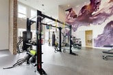 Located at 3110 Doe River Way in Matthews, NC, this expansive fitness center offers a thoughtfully designed, single-floor layout with generous open floor space spread across two distinct workout zones. The facility immediately impresses with its soaring double-height ceilings, which are complemented by oversized windows that flood the interior with abundant natural light and provide views of the surrounding neighborhood.

The design aesthetic is striking, blending contemporary and industrial elements. A dramatic large-scale abstract mural in deep purples and mauves anchors the main workout area, while an exposed brick accent wall adds warmth and texture. Durable rubber flooring throughout ensures safety and functionality across all zones.

The main strength and functional training area is outfitted with a multi-functional Octagon rig featuring TRX suspension trainers, battle ropes, a heavy bag, and kettlebells. A full dumbbell rack, weight benches, and cable machines round out the strength equipment selection. Mounted flat-screen TVs provide entertainment throughout.

A dedicated cardio zone offers spin bikes, treadmills, a stair climber, an elliptical, a rowing machine, and additional cardio equipment positioned beside floor-to-ceiling windows. A separate functional training room with open floor space, step platforms, and medicine balls provides ample room for group or individual training sessions. A hallway connects the zones, lined with large mirrors and artistic wall treatments.