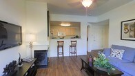 This single-floor residence showcases an open-concept design that seamlessly connects the living and kitchen areas. The living room is anchored by a sleek black fireplace and features rich hardwood flooring that extends throughout the main living space. A ceiling fan with a light fixture provides both comfort and illumination.

The kitchen is positioned behind a raised pass-through counter with bar seating for two, offering a practical and social layout ideal for casual dining or entertaining. The kitchen itself is well-appointed with light wood cabinetry, a built-in microwave, and ample counter space, all illuminated by a flush-mount ceiling light. The pass-through design maintains an open, connected feel while clearly defining the cooking area.

An elegant arched doorway adds a subtle architectural detail, providing a graceful transition between spaces. The neutral, light-toned walls throughout keep the interior feeling bright and airy. The overall ambiance is warm and inviting, combining functional design elements with comfortable, everyday living essentials.