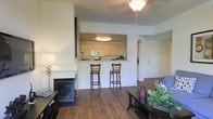 This single-floor residence showcases an open-concept design that seamlessly connects the living and kitchen areas. The living room is anchored by a sleek black fireplace and features rich hardwood flooring that extends throughout the main living space. A ceiling fan with a light fixture provides both comfort and illumination.

The kitchen is positioned behind a raised pass-through counter with bar seating for two, offering a practical and social layout ideal for casual dining or entertaining. The kitchen itself is well-appointed with light wood cabinetry, a built-in microwave, and ample counter space, all illuminated by a flush-mount ceiling light. The pass-through design maintains an open, connected feel while clearly defining the cooking area.

An elegant arched doorway adds a subtle architectural detail, providing a graceful transition between spaces. The neutral, light-toned walls throughout keep the interior feeling bright and airy. The overall ambiance is warm and inviting, combining functional design elements with comfortable, everyday living essentials.