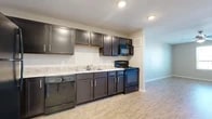 This single-floor unit offers a thoughtfully designed layout that balances functionality and comfort throughout its living spaces. The kitchen serves as a central highlight, featuring rich dark espresso-toned cabinetry with modern hardware, complemented by a full suite of black appliances including a refrigerator, range with oven, built-in microwave, and dishwasher. Light granite-style countertops provide a clean contrast against the dark cabinetry, while recessed lighting illuminates the space effectively. Light wood-style luxury flooring runs consistently throughout the unit, adding warmth and visual continuity from room to room. The bedroom is a well-proportioned space bathed in natural light from a large window with exterior views, and includes a dedicated closet with hanging storage. Neutral gray walls throughout create a calm, versatile backdrop suited to a variety of furnishing styles. The bathroom is finished with white tile flooring, a full soaking tub and shower combination, a dark-cabineted vanity with a white countertop and sink, a large wall mirror, and a multi-bulb vanity light fixture. The overall ambiance is clean, modern, and move-in ready, with consistent finishes that tie the entire space together cohesively.