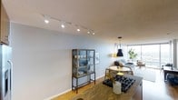 This single-floor space showcases an open-concept design that seamlessly connects the kitchen, dining, and living areas into one cohesive living environment. The kitchen features a dark granite or stone countertop with a built-in gas cooktop, offering a functional and stylish cooking space that flows naturally into the rest of the interior.

Track lighting runs along the textured ceiling, providing adjustable illumination throughout the main living area, while a sleek pendant light with a contrasting yellow-and-black shade adds a focal point above the dining area. A round white dining table paired with natural wood chairs creates an inviting setting for meals.

An industrial-style glass-front display cabinet with a metal frame serves as a standout storage piece, adding character and a touch of mixed-material design to the space. The living area is anchored by a comfortable sofa and complementary furnishings in neutral tones.

Light hardwood flooring runs throughout, contributing warmth and continuity. A large window at the far end floods the space with natural light and offers views of greenery outside, enhancing the overall sense of openness and airiness.