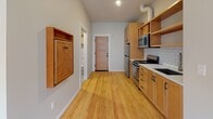 This single-floor unit combines functional design with clean, modern finishes throughout its thoughtfully laid-out spaces. Warm light hardwood flooring runs continuously throughout the unit, lending a cohesive and bright feel to every room.

The bedroom features two double-hung windows framed in dark wood trim, allowing natural light to fill the space. Dual wall-mounted mini-split units provide individual climate control, ensuring year-round comfort.

The open living and kitchen area is anchored by a full-height white cabinetry unit, offering substantial built-in storage. A wall-mounted fold-down surface adds flexible functionality. The kitchen is well-appointed with warm-toned wood cabinetry, open shelving, a white subway tile backsplash, and a full suite of stainless steel appliances including a gas range, microwave, refrigerator, and sink. A pink-toned interior door leads to the unit's entry.

The bathroom maintains a classic aesthetic with black-and-white hexagonal mosaic tile flooring, white subway tile in the shower surround, a wall-mounted porcelain basin sink, and a recessed medicine cabinet mirror. The overall ambiance of the unit is clean, practical, and move-in ready.