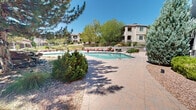 Located at 6200 Montano Plaza Dr NW in Albuquerque, New Mexico, this three-floor residential community offers a compelling blend of Southwestern architectural design and resort-style amenities set against the backdrop of the high desert landscape. The buildings feature stucco exteriors with warm earth tones, balconies, and pitched rooflines that reflect the regional aesthetic, surrounded by mature trees and native desert plantings.

At the heart of the community lies an expansive outdoor recreation area anchored by a generously sized swimming pool with crystal-clear turquoise water, complemented by a separate spa with stone-accented surrounds. The pool deck is finished with paver stonework and outfitted with red lounge chairs, providing ample space for relaxation under Albuquerque's characteristically blue skies. A shaded seating area with an umbrella offers a comfortable retreat from the sun.

The landscaping throughout the grounds is thoughtfully designed, incorporating a mix of evergreen pines, deciduous trees, ornamental shrubs, and desert-adapted plantings with decorative gravel beds. A standout water feature — a natural stone waterfall cascading over boulders — adds a tranquil, organic element to the outdoor environment. Wrought iron fencing and stone pillar accents define the perimeter, contributing to the community's polished and cohesive aesthetic. The clubhouse structure, with its large windows and stacked stone detailing, serves as a central amenity hub for the community.