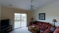 This single-floor space features a welcoming living room designed for both comfort and functionality. The room is anchored by large sliding glass doors that provide direct access to an outdoor deck or balcony, filling the space with natural light and offering views of the surrounding residential neighborhood and landscaping. The sliding doors create a seamless connection between the interior and exterior, enhancing the overall sense of openness.

The living room is furnished with a generous, deep-red upholstered sofa and matching armchair, centered around a wooden coffee table with built-in storage drawers — a practical and stylish focal point. A ceiling fan with an integrated light fixture is mounted overhead, contributing to air circulation and ambient lighting throughout the room. Neutral-toned carpet and light-colored walls create a clean, understated backdrop that makes the space feel airy and versatile.

A floor lamp positioned near a side table provides additional task lighting, while framed wall art adds a decorative touch to the otherwise minimalist décor. The overall ambiance is casual and comfortable, suited for everyday living.
