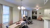 This single-story space showcases an open-concept layout that connects the dining and living areas in a cohesive, functional arrangement. The dining area is anchored by a dark wood table and illuminated by an elegant multi-arm chandelier with white glass shades, adding a refined focal point overhead. Neutral-toned walls and light carpet flooring throughout create a warm, understated backdrop that complements the varied furnishings.

Large windows along the exterior wall allow natural light to filter into the dining space, dressed with floor-length curtains featuring subtle orange accents. The adjoining living area is outfitted with a ceiling fan and light fixture, contributing to comfortable air circulation. A dark sofa, a floral-patterned accent chair, and a side table define the seating zone, while built-in shelving visible in the background provides practical storage and display space.

The transition between the dining and living areas is seamless, enhancing the sense of openness throughout. The overall ambiance is casual yet intentional, with layered lighting, decorative accents, and a mix of textures contributing to a lived-in, welcoming atmosphere suited for everyday use and entertaining.