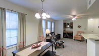 This single-story space showcases an open-concept layout that connects the dining and living areas in a cohesive, functional arrangement. The dining area is anchored by a dark wood table and illuminated by an elegant multi-arm chandelier with white glass shades, adding a refined focal point overhead. Neutral-toned walls and light carpet flooring throughout create a warm, understated backdrop that complements the varied furnishings.

Large windows along the exterior wall allow natural light to filter into the dining space, dressed with floor-length curtains featuring subtle orange accents. The adjoining living area is outfitted with a ceiling fan and light fixture, contributing to comfortable air circulation. A dark sofa, a floral-patterned accent chair, and a side table define the seating zone, while built-in shelving visible in the background provides practical storage and display space.

The transition between the dining and living areas is seamless, enhancing the sense of openness throughout. The overall ambiance is casual yet intentional, with layered lighting, decorative accents, and a mix of textures contributing to a lived-in, welcoming atmosphere suited for everyday use and entertaining.