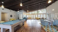 Located at 603 Plum Nearly Ln in Wilmington, NC, this single-story community clubhouse is a well-appointed gathering space designed for both relaxation and socializing. The architecture is immediately impressive, characterized by soaring exposed ceilings with dark structural beams and pendant lighting that lend the space an open, industrial-chic aesthetic. Rich hardwood flooring runs throughout, tying the various zones together cohesively.

The main hall is generously proportioned, accommodating a full-size billiards table as a centerpiece recreational feature. Adjacent to this is a comfortable lounge area furnished with plush seating — including soft yellow armchairs and gray accent chairs — arranged atop colorful geometric-patterned area rugs. A wall-mounted flat-screen television anchors one end of the lounge, making it suitable for communal viewing.

Floor-to-ceiling glass walls and large double-hung windows line one side of the space, flooding the interior with abundant natural light and offering serene views of the surrounding greenery. French doors provide direct access to the outdoors, enhancing the connection between interior and exterior.

A kitchenette area is neatly integrated into the layout, equipped with gray cabinetry, dark countertops, a stainless steel refrigerator, and a coffee station — ideal for hosting events or casual gatherings. A dedicated hallway leads to additional utility rooms. Overall, the space presents a bright, modern, and welcoming environment.