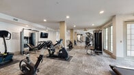 This expansive fitness center is thoughtfully designed to support a full spectrum of workout routines. The single-floor layout is organized into distinct zones, offering an impressive array of both cardio and strength training equipment. A dedicated cardio section lines the perimeter, featuring multiple ProMAXIMA treadmills positioned in front of large windows that invite natural light and views of the outdoors, including a pool area visible from one end of the facility. Additional cardio options include stationary bikes, elliptical trainers, and a rowing machine. The strength training area is equally well-appointed, with cable machines, a functional training rig complete with a punching bag and battle ropes, a trampoline, TRX suspension straps, and a comprehensive dumbbell rack with an overhead mounted flat-screen TV. Structural columns clad in warm wood-toned paneling add an architectural detail that breaks up the open floor plan while contributing to the contemporary aesthetic. Recessed ceiling lighting and decorative gold ceiling fans ensure consistent illumination throughout. The flooring consists of durable, multi-toned carpet tiles. Mirrored walls in select areas visually expand the space, while large windows throughout maintain a bright and energizing atmosphere well-suited to an active fitness environment.