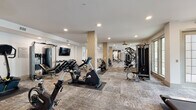This expansive fitness center is thoughtfully designed to support a full spectrum of workout routines. The single-floor layout is organized into distinct zones, offering an impressive array of both cardio and strength training equipment. A dedicated cardio section lines the perimeter, featuring multiple ProMAXIMA treadmills positioned in front of large windows that invite natural light and views of the outdoors, including a pool area visible from one end of the facility. Additional cardio options include stationary bikes, elliptical trainers, and a rowing machine. The strength training area is equally well-appointed, with cable machines, a functional training rig complete with a punching bag and battle ropes, a trampoline, TRX suspension straps, and a comprehensive dumbbell rack with an overhead mounted flat-screen TV. Structural columns clad in warm wood-toned paneling add an architectural detail that breaks up the open floor plan while contributing to the contemporary aesthetic. Recessed ceiling lighting and decorative gold ceiling fans ensure consistent illumination throughout. The flooring consists of durable, multi-toned carpet tiles. Mirrored walls in select areas visually expand the space, while large windows throughout maintain a bright and energizing atmosphere well-suited to an active fitness environment.