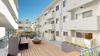 This multi-story residential community centers around a vibrant, well-appointed courtyard that serves as the heart of the property. The exterior architecture is characterized by clean white stucco facades, protruding balconies, and decorative lattice screen panels that add a mid-century modern aesthetic to the overall design.

The courtyard is a standout feature, showcasing a generously sized concrete plaza adorned with a bold geometric pattern in soft tones of teal, sage green, and cream. Large, sculptural dark bowl planters filled with young ornamental trees punctuate the open space, adding greenery and visual interest. An outdoor dining area with a wood table and chairs sits beneath a pergola, offering a shaded gathering space bathed in natural light.

An elevated wood deck platform, accessible via wide timber steps with metal railings, provides a secondary outdoor lounge area complete with a central fire bowl. The deck is flanked by raised planter beds with ornamental grasses, contributing to the landscaped feel of the complex.

Open-air walkways with teal-painted metal railings connect the building levels, while accent doors in vibrant green add pops of color throughout. The individual unit patios offer private outdoor spaces that open directly onto the colorfully patterned courtyard below, ensuring residents enjoy both community and privacy within this sun-filled setting.