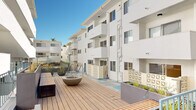 This multi-story residential community centers around a vibrant, well-appointed courtyard that serves as the heart of the property. The exterior architecture is characterized by clean white stucco facades, protruding balconies, and decorative lattice screen panels that add a mid-century modern aesthetic to the overall design.

The courtyard is a standout feature, showcasing a generously sized concrete plaza adorned with a bold geometric pattern in soft tones of teal, sage green, and cream. Large, sculptural dark bowl planters filled with young ornamental trees punctuate the open space, adding greenery and visual interest. An outdoor dining area with a wood table and chairs sits beneath a pergola, offering a shaded gathering space bathed in natural light.

An elevated wood deck platform, accessible via wide timber steps with metal railings, provides a secondary outdoor lounge area complete with a central fire bowl. The deck is flanked by raised planter beds with ornamental grasses, contributing to the landscaped feel of the complex.

Open-air walkways with teal-painted metal railings connect the building levels, while accent doors in vibrant green add pops of color throughout. The individual unit patios offer private outdoor spaces that open directly onto the colorfully patterned courtyard below, ensuring residents enjoy both community and privacy within this sun-filled setting.
