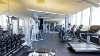 This fitness center is thoughtfully designed to accommodate a comprehensive workout experience. The space is lined with floor-to-ceiling windows along one side, flooding the interior with natural light and offering open views of the surrounding area. The expansive layout is divided into distinct zones, with cardio equipment — including ellipticals, treadmills, and similar machines — arranged in a row along the windowed wall, allowing users to exercise while enjoying the view. On the opposite side, a well-appointed strength training area features cable machines, weight benches, and various resistance equipment. Full-length mirrors are mounted along the walls, enhancing the sense of depth and allowing users to monitor their form. The flooring consists of durable dark rubber matting, practical for both heavy equipment and high-traffic use. Overhead, recessed and linear lighting supplements the natural light, ensuring consistent illumination throughout. Individual screens are mounted on select cardio machines for personal entertainment. The overall ambiance is sleek, modern, and functional, reflecting a professional-grade facility designed to support a wide range of fitness goals.