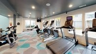 This contemporary fitness center is thoughtfully designed to support a full range of workout routines across two distinct zones. The cardio area features a bold geometric-patterned carpet in teal, beige, and gray tones, and is outfitted with treadmills, ellipticals, stationary bikes, and a rowing machine. A dark-painted ceiling with recessed lighting adds a sleek, modern contrast, while large windows along the perimeter flood the space with natural light and offer views of the surrounding outdoor environment. Wall-mounted televisions provide entertainment throughout.

The strength and functional training zone is distinguished by soaring ceilings and clean white walls accented by a large motivational graphic mural featuring fitness-themed typography. This area includes free weights, a weight bench, resistance equipment, and racks stocked with medicine balls, resistance bands, and exercise mats. An interactive fitness display screen serves as a digital training resource. Light wood-look flooring anchors this zone with a warm, clean aesthetic. A towel station with open cubbies adds a convenient amenity touch. The space is finished with modern linear pendant lighting and glass double doors that enhance openness and connectivity throughout the facility.