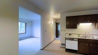 This single-floor space presents a straightforward, functional layout with a clean, neutral aesthetic. The kitchen area is equipped with a white range and dark wood cabinetry overhead, providing a classic contrast against the light-colored walls and flooring. A wall-mounted light fixture adds a soft, ambient glow to the space.

A wide arched opening connects the kitchen to the adjacent living area, which features light carpeting, a baseboard heater, and a window that allows natural light to filter in, offering a view of surrounding greenery. The open sightline between these two areas enhances the sense of space and connectivity.

A hallway visible from the kitchen leads further into the unit, with a doorway at its end hinting at additional rooms beyond. The corridor is well-lit and maintains the same neutral wall tones seen throughout, contributing to a cohesive and consistent interior palette.

Overall, the space features a practical, no-frills design with white walls, light carpeting, and modest built-in elements. The layout prioritizes ease of movement and functional living across its single floor.