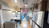 This one-of-a-kind unit at Germantown Mill Lofts is housed within a converted train car, offering a distinctive living experience that preserves the vehicle's original structural character while incorporating modern interior updates. The elongated, single-floor layout follows the natural form of the rail car, with warm hardwood floors running the full length of the space and contributing a sense of continuity throughout.

The kitchen is efficiently designed, featuring stainless steel appliances including a refrigerator, electric range, microwave, and a single-basin sink set into a wood-toned countertop. Cabinetry above and below provides functional storage, and a small window above the sink brings in natural light. Adjacent to the kitchen, a cozy dining nook features built-in booth-style seating in gray upholstery arranged around a compact table, with windows offering views of the surrounding environment.

The corridor-style hallway connects the various sections of the space, with exposed conduit running along the ceiling — a nod to the industrial origins of the structure. Separate enclosed rooms branch off the main corridor, offering defined private areas. The open end room, featuring a door with a window, provides additional flexible space. A wall-mounted mini-split unit handles climate control throughout.