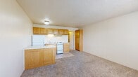 This single-floor unit offers a practical, open-concept layout where the kitchen and living area flow seamlessly together. The kitchen is outfitted with warm oak cabinetry both above and below the counters, providing ample storage space. A peninsula with a light-toned countertop extends into the living area, creating a natural divide between the two spaces while maintaining an open feel. White appliances include a refrigerator, electric range, and a built-in range hood. The kitchen floor is finished in light-toned ceramic tile, which transitions into wall-to-wall carpeting throughout the living area. A tall standalone oak cabinet near the kitchen adds additional storage. The living area is generously proportioned with neutral walls and a textured ceiling, creating a clean, understated ambiance. A sliding window allows natural light into the space, and a wall-mounted air conditioning unit provides climate control. A doorway leads to a bathroom area that includes a vanity with an oak cabinet base and an adjacent closet with a hanging rod and shelving, offering convenient storage. The overall design is functional and straightforward, with consistent warm wood tones throughout.