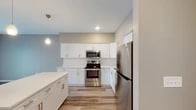 This single-floor residence offers a thoughtfully designed layout that balances modern style with everyday functionality. The kitchen serves as a standout feature, showcasing crisp white shaker cabinetry, light-toned countertops, and a full suite of stainless steel appliances including a range with microwave above and a full-size refrigerator. A large kitchen island provides additional prep and dining space, while pendant lighting adds a contemporary touch overhead. Wood-look flooring runs throughout the kitchen and main living areas, contributing to a clean, cohesive aesthetic. The living area transitions seamlessly from the kitchen and features neutral-toned walls with plush carpeting underfoot, creating a comfortable and inviting atmosphere. Bedrooms are carpeted and fitted with ceiling fans, and natural light enters through window openings fitted with blinds. A dedicated in-unit laundry room accommodates a full-size, top-loading washer and dryer side by side, complete with overhead wire shelving for added storage convenience. The bathroom features a wide vanity with a solid white countertop, an integrated sink, a large mirror, and a glass-enclosed shower stall with tiled surround. Throughout the space, neutral warm-gray wall tones and consistent finishes create a unified, modern residential environment.