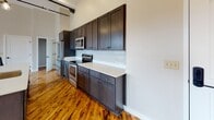 Located in Hornell, NY, this two-floor loft blends industrial character with modern updates across a thoughtfully designed layout. The main living area makes a strong impression with soaring ceilings, exposed silver ductwork, and dark steel beams that contrast beautifully against white walls. Original hardwood floors with natural variation run throughout, adding warmth and texture to the open-concept space. Large windows with dark roller shades flood the area with natural light, while pendant lighting in varied styles reinforces the industrial aesthetic.

The kitchen is well-appointed with dark espresso shaker cabinets extending to the ceiling, white quartz countertops, and a full suite of stainless steel appliances. A kitchen island with an undermount sink provides additional prep space and serves as a natural divider between the kitchen and living areas.

The bedrooms offer a quieter, more refined atmosphere, with painted brick accent walls and sunlight streaming across the hardwood floors. The bathrooms are distinctly finished — one features large-format wood-look tile flooring and a spacious glass-enclosed walk-in shower with floor-to-ceiling tile surround, while another showcases an angled ceiling with an exposed dark beam, adding architectural interest. Both bathrooms include quartz vanity surfaces and brushed nickel fixtures throughout.