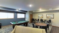 This single-floor common amenity lounge is thoughtfully designed to serve as a social and recreational hub. The space is anchored by a full-sized pool table with a green felt surface, positioned beneath recessed ceiling lighting that ensures excellent visibility for gameplay. The room features a combination of blue-gray and neutral beige wall tones, creating a relaxed yet polished atmosphere.

Seating options are varied and plentiful. High-top bar tables paired with black cushioned stools line the perimeter, offering elevated seating ideal for casual conversation or spectating. A round table with matching dark chairs provides additional gathering space, while a light-colored upholstered sofa at the forefront of the room adds a comfortable lounge element.

Natural light filters into the space through windows dressed with simple blinds, supplemented by recessed overhead lighting throughout. The patterned low-pile carpet adds a layer of texture and visual interest underfoot. A framed artwork piece adorns one wall, contributing a decorative touch. The overall ambiance is casual and inviting, well-suited for group socializing and leisure activities.
