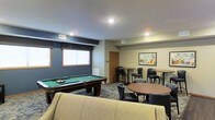 This single-floor common amenity lounge is thoughtfully designed to serve as a social and recreational hub. The space is anchored by a full-sized pool table with a green felt surface, positioned beneath recessed ceiling lighting that ensures excellent visibility for gameplay. The room features a combination of blue-gray and neutral beige wall tones, creating a relaxed yet polished atmosphere.

Seating options are varied and plentiful. High-top bar tables paired with black cushioned stools line the perimeter, offering elevated seating ideal for casual conversation or spectating. A round table with matching dark chairs provides additional gathering space, while a light-colored upholstered sofa at the forefront of the room adds a comfortable lounge element.

Natural light filters into the space through windows dressed with simple blinds, supplemented by recessed overhead lighting throughout. The patterned low-pile carpet adds a layer of texture and visual interest underfoot. A framed artwork piece adorns one wall, contributing a decorative touch. The overall ambiance is casual and inviting, well-suited for group socializing and leisure activities.