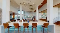 This expansive single-floor amenity space showcases a bold mid-century modern aesthetic, seamlessly blending architectural elegance with vibrant, curated design. Soaring double-height ceilings flood the open-plan interior with natural light through floor-to-ceiling glazing, offering views of the surrounding landscaped exterior. Polished terrazzo floors extend throughout, lending a clean, luminous foundation to the space.

At the heart of the main lounge, dramatic wire-frame pendant light fixtures serve as sculptural focal points overhead, complementing the warm wood ceiling accents. Multiple seating areas are thoughtfully arranged, featuring classic mid-century furnishings including Eames lounge chairs, egg chairs, and plush sofas in earthy and neutral tones, anchored by boldly patterned area rugs.

A long communal bar counter with teal geometric tile detailing and warm wood barstools provides a social gathering point, while a separate lounge room features a striking suspended fireplace, patterned tile flooring, and rich orange upholstered seating beneath a slatted wood ceiling. Decorative breeze-block screens and textured accent walls add depth and visual interest. Glass-walled private offices or meeting rooms are accessible off the main corridor. A built-in bookshelf and stepped platform area with warm wood finishes complete this thoughtfully designed, community-oriented space.